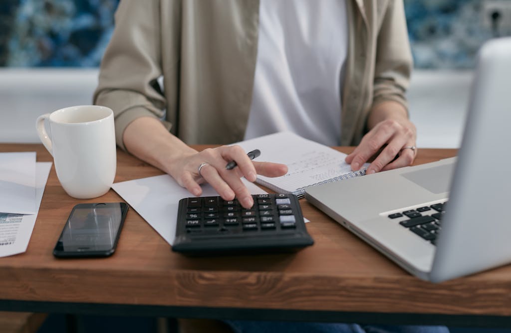A person using a calculator and laptop while taking notes at a wooden desk.
